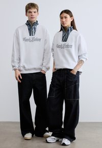 Δύο άτομα φορούν λευκά φούτερ με γιακά με σχέδιο και σκούρα τζιν με φαρδιά πόδια. Σημαντικές λεπτομέρειες περιλαμβάνουν εκτυπωμένη επωνυμία και αθλητικά παπούτσια.