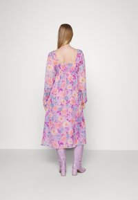 Vestido longo, floral em roxo pastel com flores coloridas, apresentando costas quadradas e cintura franzida. Combinado com botas roxas claras.