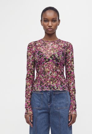 Femme portant un haut ajusté à manches longues, transparent, à motifs floraux roses, violets et beiges, associé à un pantalon en denim bleu avec de grandes poches avant.