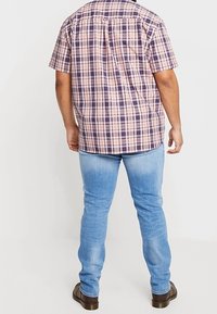 Adulto che indossa una camicia a maniche corte a quadri viola e rosa, jeans azzurri chiari e scarpe marroni, in piedi con la schiena rivolta verso la fotocamera.