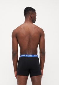 Schwarze Boxershorts mit einem blauen Bund, auf dem der Text "BJÖRN BORG" steht. Der Stoff wirkt glatt; das Design ist körpernah geschnitten und hat eine mittellange Passform.