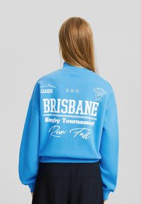 Persona con cabello largo que lleva una sudadera azul con texto blanco en la espalda que dice "Brisbane Rugby Tournament Run Fast".