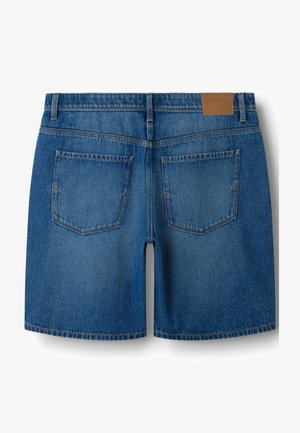 Plave traper hlače kratkih nogavica s dva stražnja džepa, petljama za remen i smeđom kožnom zakrpom na pojasu, prikazane straga.