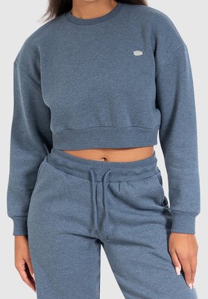 Kobieta w niebieskiej krótkiej bluzie i dopasowanych, wysokich spodniach dresowych ze sznurkiem na jasnym tle.