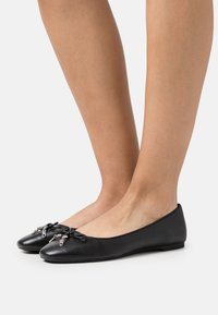 Ballerine in pelle nera con punta tonda, caratterizzate da un accento a fiocco e piccoli dettagli metallici sulla parte anteriore. Texture liscia, suola piatta.