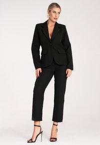 Czarna marynarka zapinana na jeden guzik z wykładanymi klapami, z dwiema przednimi kieszeniami, w połączeniu z dopasowanymi czarnymi spodniami i sandałami na obcasie.