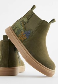 Cizme de gleznă din suede verde, cu talpă din cauciuc bej, având un design cu dinozauri și frunze pe lateral, laterale elastice și bucle de tragere.