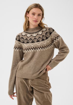 Kvinde iført en beige mønstret striktrøje og løse brune kordfløjlsbukser, poserende med den ene hånd på hoften mod en hvid baggrund.