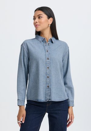 Junge Frau mit langen dunklen Haaren, die ein hellblaues, geknöpftes Jeanshemd und dunkelblaue Jeans trägt und vor weißem Hintergrund steht.
