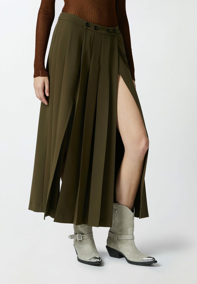 La femme porte une jupe plissée vert olive avec une fente haute, un haut marron à manches longues et des bottes de cow-boy gris clair avec des pointes métalliques.