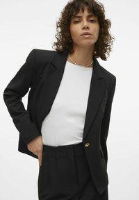 Černý blazer se strukturovanými rameny, s klopami a jediným zlatým tlačítkem. Nošený přes bílou košili, doplněný o přizpůsobené černé kalhoty.