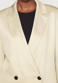 Blazer de doble botonadura color beige claro con textura suave, solapas afiladas y botones negros, superpuesto a una blusa negra.