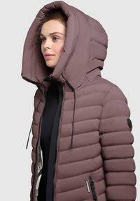 Žena nosi ljubičastu puffer jaknu s velikim kapuljačom i crnim zatvaračem, okrenuta lijevo na svijetlosivoj pozadini.