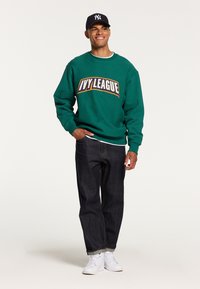 Felpa verde con la scritta "IVY LEAGUE" stampata in caratteri grassetto, abbinata a jeans di denim scuro e sneakers bianche, completata da un cappellino nero.