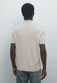 Uomo con pelle scura e capelli ricci, che indossa una polo beige a maniche corte e jeans blu, in piedi di fronte a una parete grigia chiara.