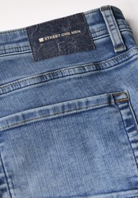 Jeans de denim azul claro con una superficie texturizada y costuras naranjas en contraste, que presentan una etiqueta negra marcada "STREET ONE MEN" en la cintura.