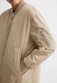 Licht beige bomberjack met een gladde textuur. Voorzien van een ritssluiting aan de voorkant, geribbelde kraag en een ritszak op de linker mouw.