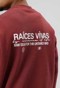 Sweat-shirt bordeaux avec texte blanc au dos, affichant "RAÍCES VIVAS" et d'autres graphiques. Tissu en coton, col rond.