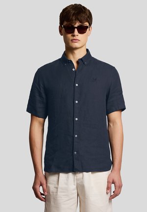 Ung mand iført mørk marineblå kortærmet skjorte med knapper, beige bukser og mørke solbriller, stående mod en ensartet lys grå baggrund.