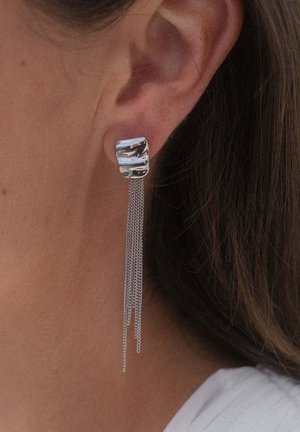 Silberner geometrischer Stecker-Ohrring mit mehreren filigranen Ketten, getragen am linken Ohr bei dunklem Haar und heller sichtbarer Haut.