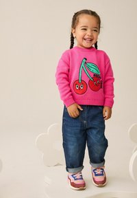 Bambino sorridente con capelli intrecciati, che indossa un maglione rosa acceso con ciliegie, jeans blu e sneakers colorate, in piedi su una piattaforma a forma di fiore bianco.
