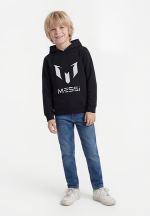 Lyshåret dreng står iført sort Messi-hoodie, blå jeans og hvide sneakers mod en ensfarvet hvid baggrund.