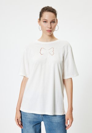 Camiseta blanca de algodón con cuello redondo, mangas cortas y un detalle de lazo bordado en el centro. Combinada con unos vaqueros de mezclilla azul claro.