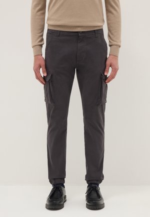 Man draagt donkergrijze cargobroek met elastische boorden, beige top met lange mouwen en zwarte veterschoenen, staand tegen een effen achtergrond.