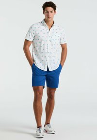 Witte kortebloemshirt met een blauw palmbomenpatroon, gecombineerd met blauwe shorts. Model draagt witte sneakers met rode accenten.