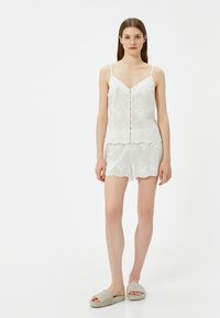 Valkoisilla kukkakuviolla varustettu toppi, jossa on napit, yhdistettynä matching-shortsien kanssa. Pehmeä kangas ja aaltoilevat reunat. Kevyt ja hengittävä muotoilu.