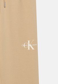 Bež jogger hlače od mekog materijala s bijelim logom "CK" i natpisom "Calvin Klein Jeans" s lijeve strane.