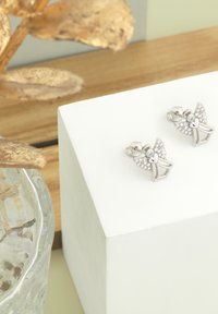 Silverfärgade ängelformade örhängen med ett hjärtdesign och dekorerade med klara ädelstenar. Visas på en vit pedestal med träbakgrund.