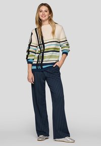 Bunter Strickpullover mit horizontalen Streifen in Creme, Blau, Grün und Schwarz, kombiniert mit weiten, navyblauen gestreiften Hosen und silbernen Schuhen.