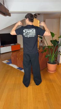 Juoda marškinėliai su baltu užrašu „STANCE PLUMBING SERVICE“ ant nugaros, derinami su laisvomis juodomis kelnėmis. Vidaus aplinka su augalais.