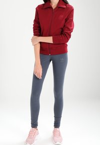 Kvinna som har på sig en röd zip-up jacka, grå Adidas leggings och rosa sneakers, står med ena armen korsad över den andra.