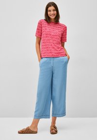 T-shirt rose à manches courtes avec un motif abstrait rouge, associé à des culottes en denim large bleu clair et des sandales marron à pois blancs.