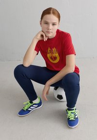 Noor punaste juustega noor inimene kannab punast Hispaania T-särki, tumesiniseid pükse ja siniseid spordijalatsid, istub jalgpallil siseruumides.