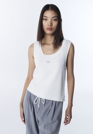 Jonge vrouw met steil donker haar, gekleed in een witte, mouwloze gebreide top en losse grijze broek, tegen een effen witte achtergrond.