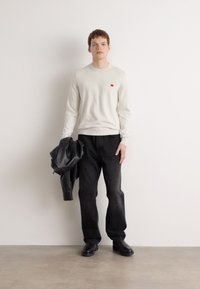 Un pull gris clair avec un petit logo rouge, un jean noir et une veste en cuir noire drapée sur un bras. Le mannequin porte des chaussures noires sur un fond neutre.