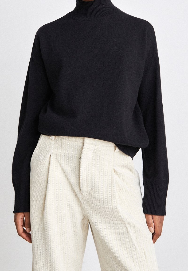 Personne portant un pull à col roulé noir rentré dans un pantalon texturé crème à taille haute avec des plis.