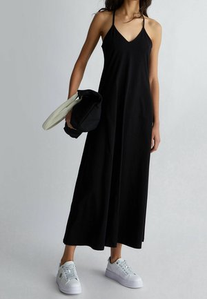 Mujer que lleva un vestido maxi negro sin mangas y zapatillas blancas, sosteniendo una chaqueta negra y blanca doblada en una mano.