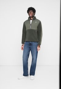 Olive green fleece pullover with a zippered collar, contrasting chest panel, paired with blue jeans and white sneakers.