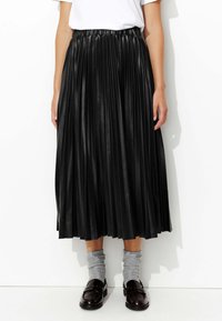 Jupe plissée noire en simili cuir avec une taille élastique. Elle est de longueur longue et est accompagnée de chaussettes grises et de mocassins.