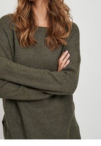 Femme portant un pull en maille vert olive à manches longues, bras croisés, avec des cheveux bruns ondulés mi-longs visibles sur un fond uni.