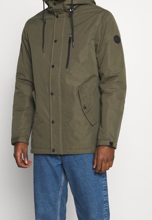 Homme portant une veste d'hiver à capuche de couleur olive avec des boutons-pression noirs et une poche zippée, assortie à un jean en denim bleu.