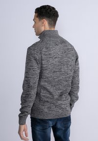 Maglione grigio lavorato a maglia con colletto alto, caratterizzato da un motivo testurizzato, polsini e orlo a coste, indossato con jeans blu scuri.