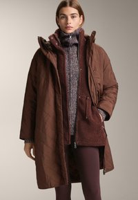 Hnedá prešívaná parka s kapucňou na textúrovanom bordovom a fialovom svetri. Obsahuje predný zips, vrecká a voľný strih.