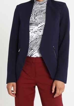 Persoon draagt een marineblauw colbert met ritszakken, een wit en zwart top met abstract patroon en donkerrode broek.