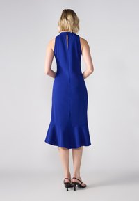 Blaues, ärmelloses Kleid mit ausgestelltem Saum, das einen Schlüsselrücken hat und eine glatte, figurbetonte Silhouette bietet. Getragen mit schwarzen Riemchen-High Heels.