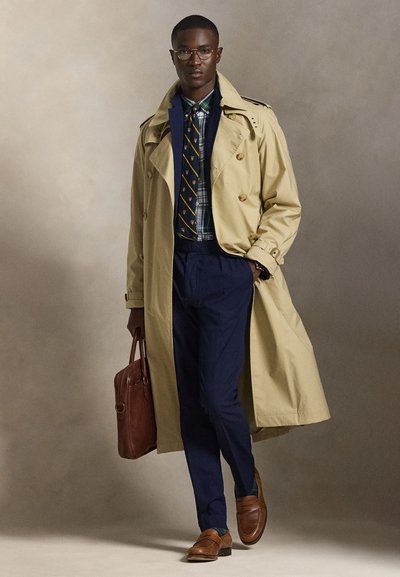 Άνδρας που φοράει μπεζ παλτό τρεντζκότ, μπλε κοστούμι, καρό πουκάμισο, γραμμωτή γραβάτα, καφέ δερμάτινα παπούτσια, κρατώντας καφέ δερμάτινο χαρτοφύλακα, περπατώντας προς τα εμπρός.